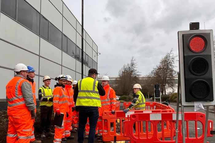 Group of construction workers in high-visibility clothing and helmets standing near a building, discussing around a work site barricaded with orange barriers. A traffic light shows a red signal.