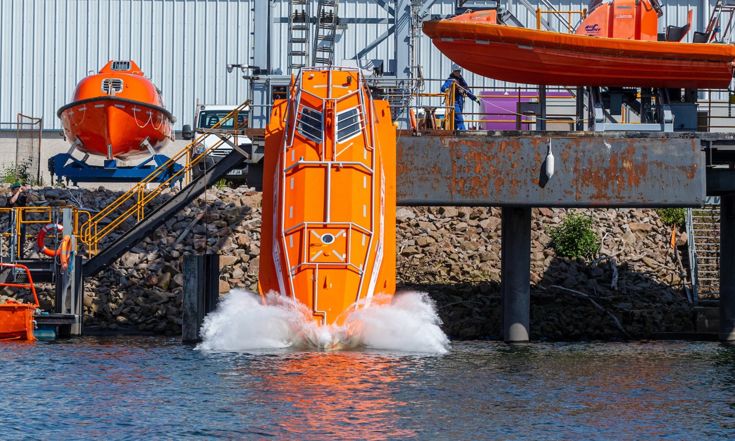 A bright orange lifeboat is being launched from a dock into the water, creating a splash. There are additional lifeboats and industrial structures in the background.