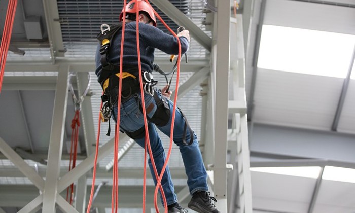 A person wearing safety gear and a red helmet is suspended by ropes, climbing on a metal structure inside an industrial facility.