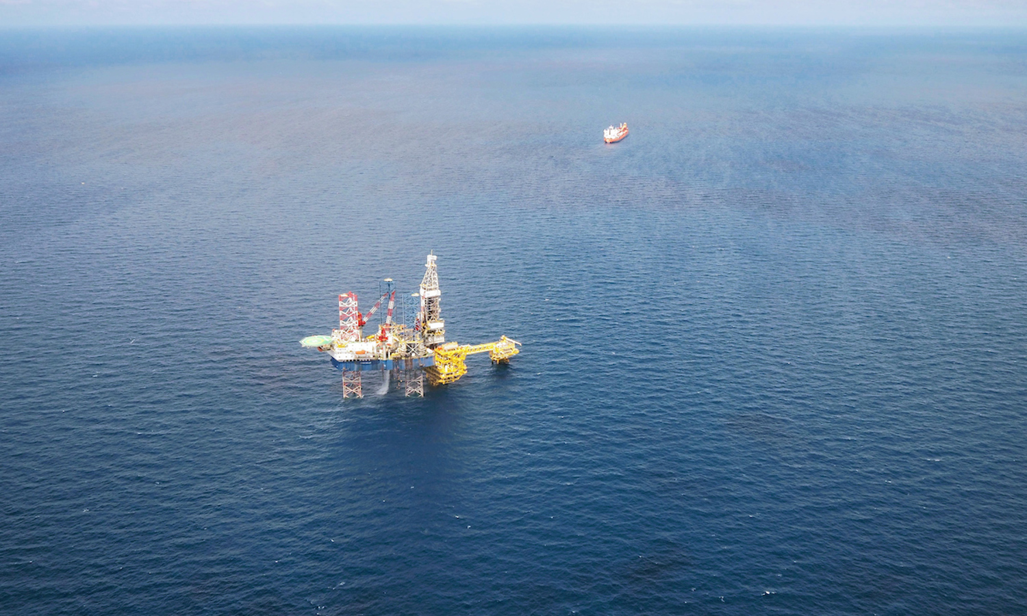 Aerial view of an offshore oil platform in the ocean, with a distant ship on the horizon under a partly cloudy sky, subtly illustrating nature's delicate balance amid the relentless training of industry.