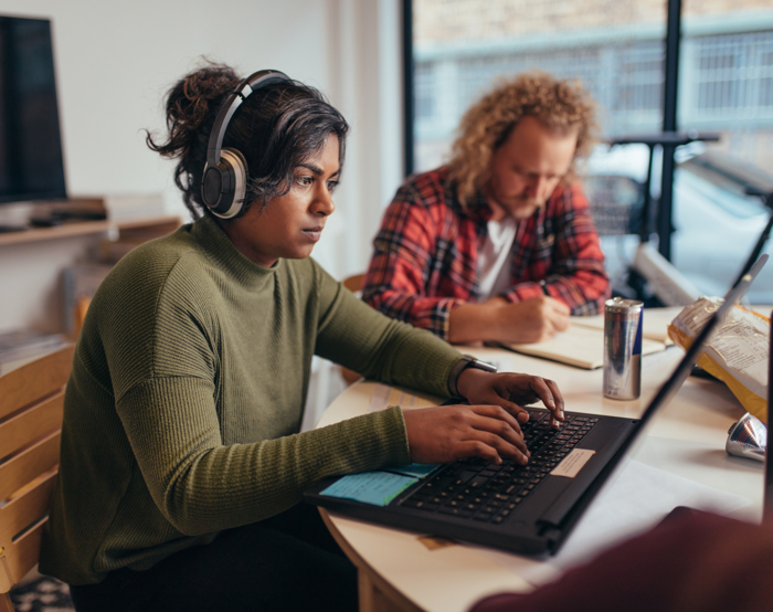 A person with headphones works on a laptop at a table, focused on training modules, while another person writes in the background.