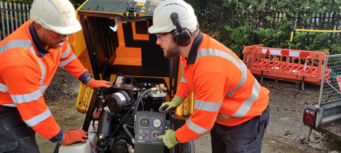 Two workers in orange high-visibility jackets and helmets undergo training on site, operating equipment with precision. One expertly handles the control panel while the other keenly observes. Safety barriers and greenery frame the scene, ensuring both safety and a touch of nature.