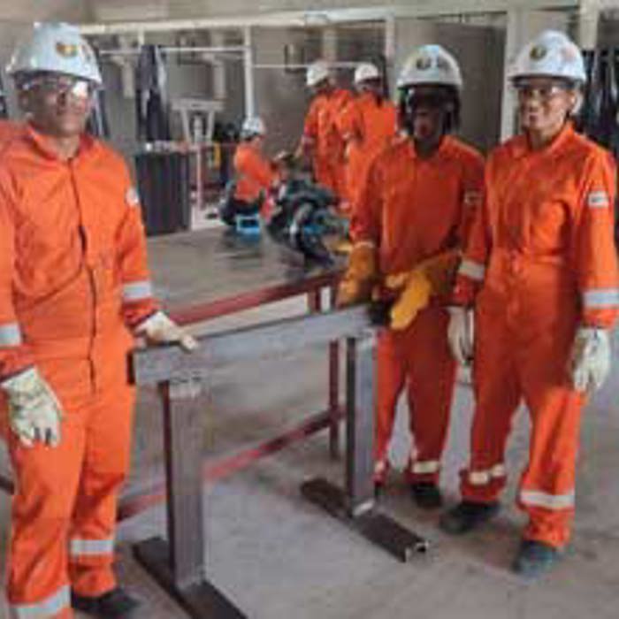 A group of workers in orange coveralls and white helmets stand around a metal structure in an industrial workshop.