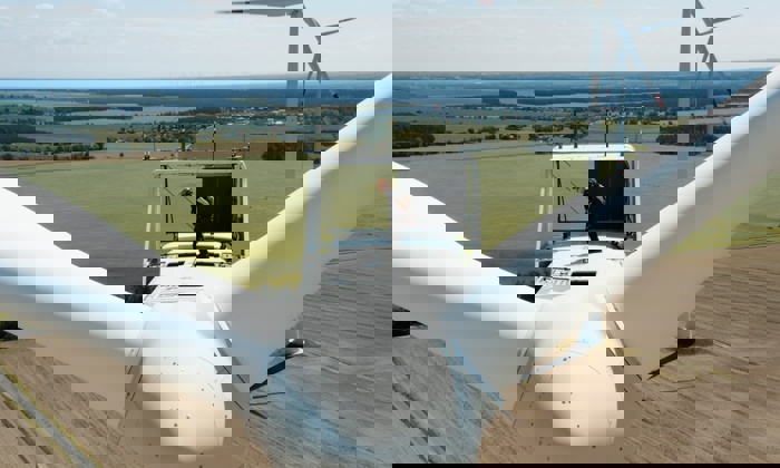 A technician is standing on top of a wind turbine in a rural area, performing maintenance. The surrounding landscape consists of expansive fields and distant tree lines.