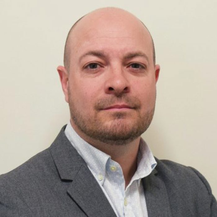 A bald man with a beard is wearing a grey suit and light-colored shirt, standing against a plain background.