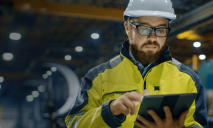 Man in high-visibility jacket and hard hat uses a tablet in an industrial setting.
