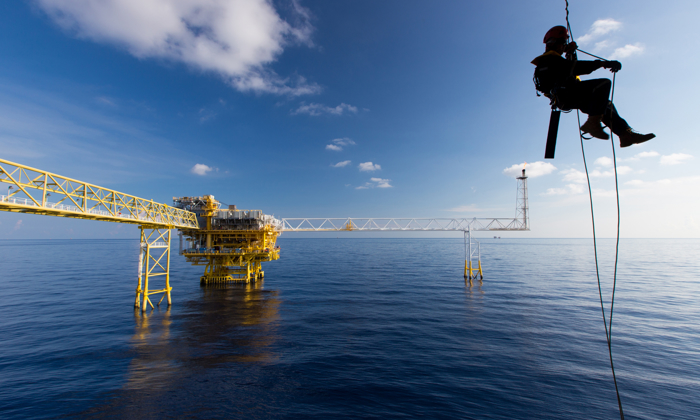 A worker, undergoing training, is suspended by a safety rope above the ocean, with an offshore oil platform in the background under a blue sky with scattered clouds.