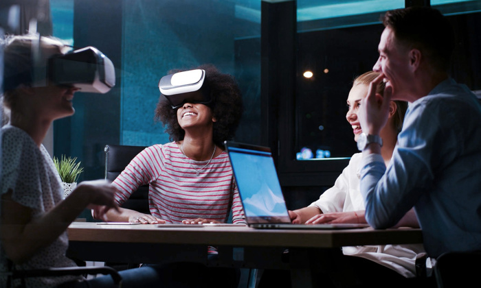Four people sit at a table in a dimly lit room, two wearing virtual reality headsets, laughing and appearing to enjoy the experience. A laptop is open on the table.