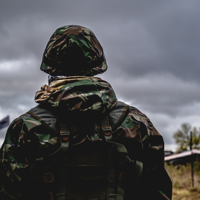 A person in camouflage military gear stands outdoors, possibly engaging in training exercises, under a cloudy sky.