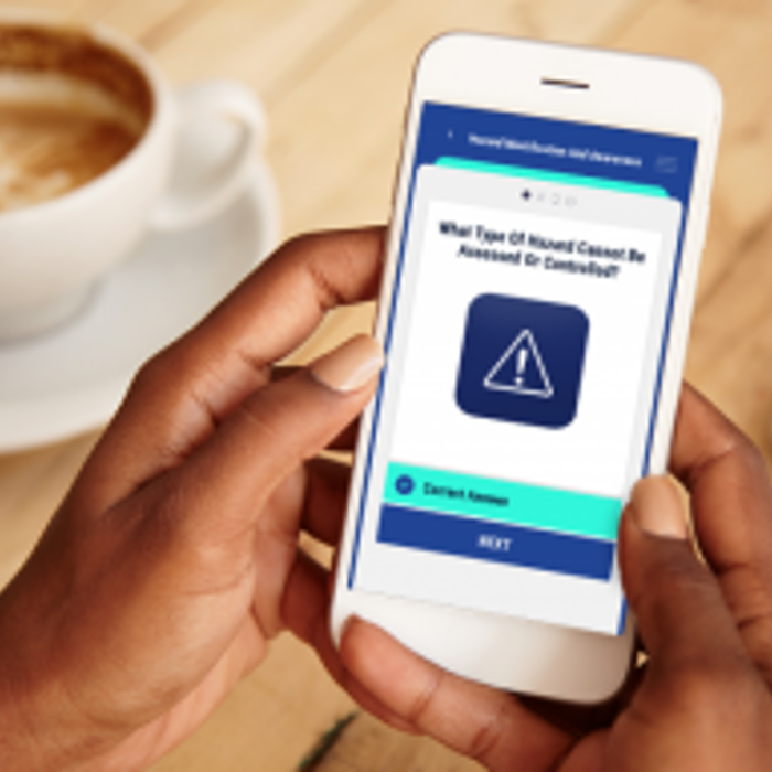 A person holds a smartphone displaying a question with a warning symbol on a quiz app. A cup of coffee and a laptop are on the wooden table in the background.