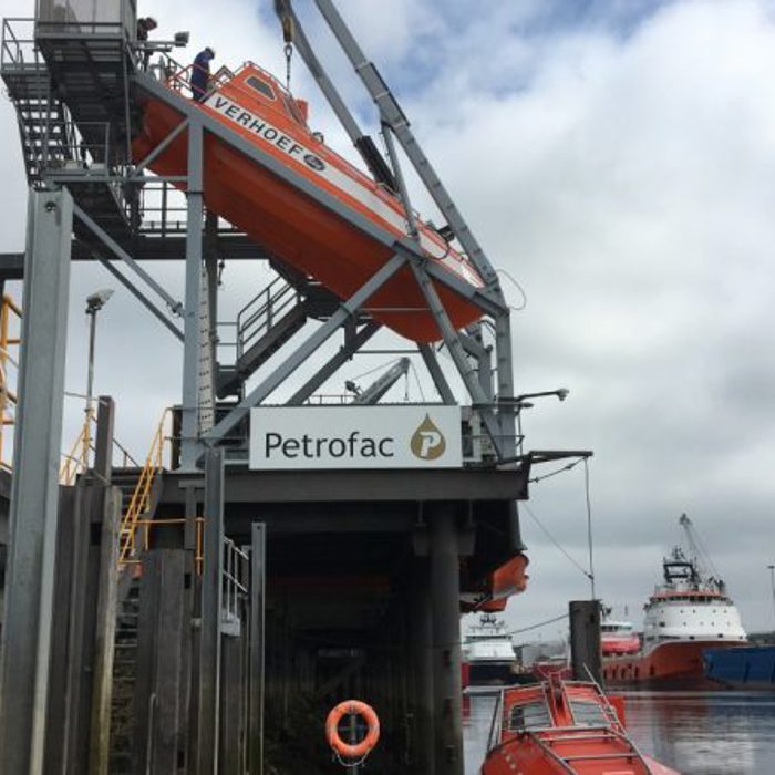 An orange lifeboat is suspended on a launching mechanism above the water at a Petrofac facility, with two ships docked in the background.