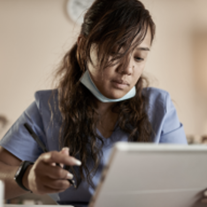 A person in a blue medical uniform, wearing a mask under their chin, is looking at a tablet and writing with a stylus.