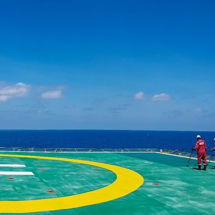 Three workers in red uniforms stand on a large helipad near the ocean under a clear blue sky.