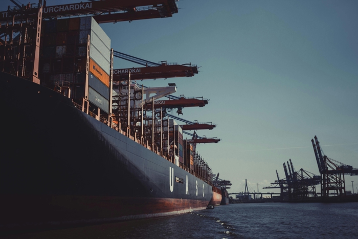 Container ship docked at a port with large cranes overhead, set against a clear sky and calm water.