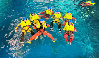 Seven individuals in bright yellow flotation suits and blue helmets float in a swimming pool, practicing water safety or rescue training techniques as precise as those performed by the military.