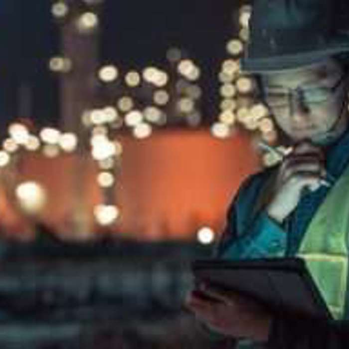 A worker wearing a hard hat and reflective vest uses a tablet in an industrial area at night. Illuminated structures in the background.