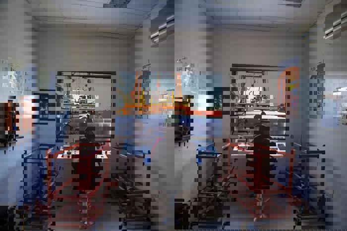 Interior of a control room with a person seated at a console, monitoring a crane lifting a container outside. Red equipment frames flank the room, and informational posters are on the walls.