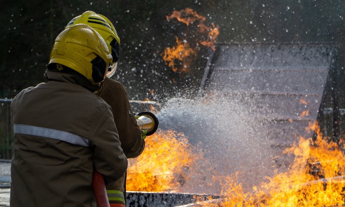 Two firefighters in protective gear aim a hose to extinguish a fire, with flames and smoke billowing around.