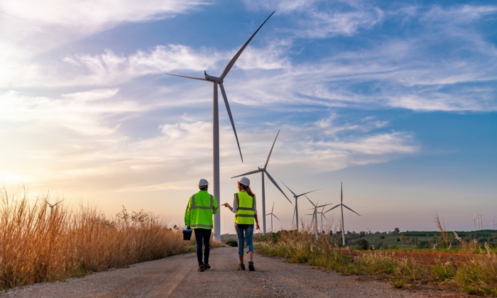 Two people in safety vests walk on a path surrounded by wind turbines under a blue sky, participating in valuable training on renewable energy.