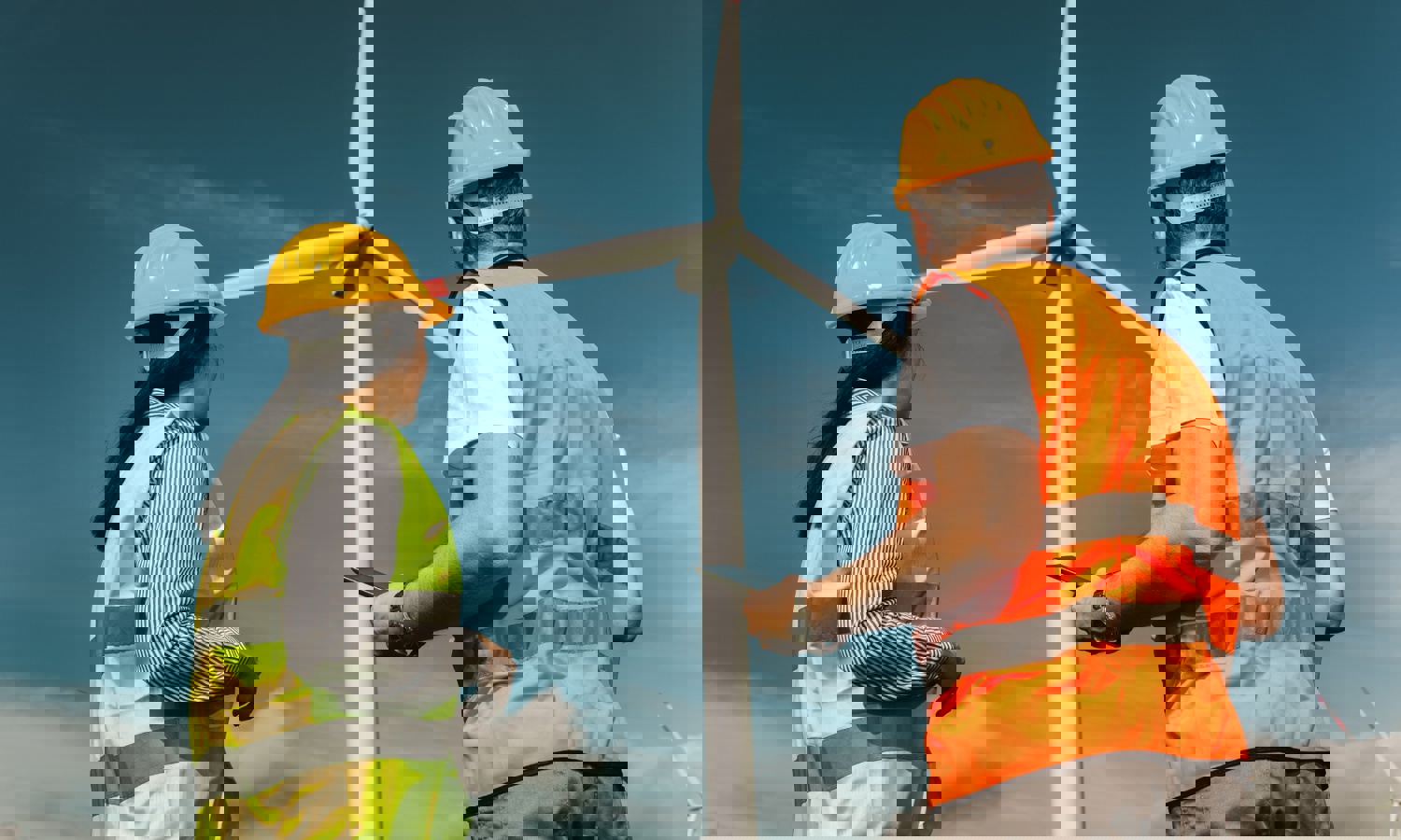 Two workers wearing hard hats and reflective vests stand near a large wind turbine, observing and possibly inspecting it under a clear blue sky.