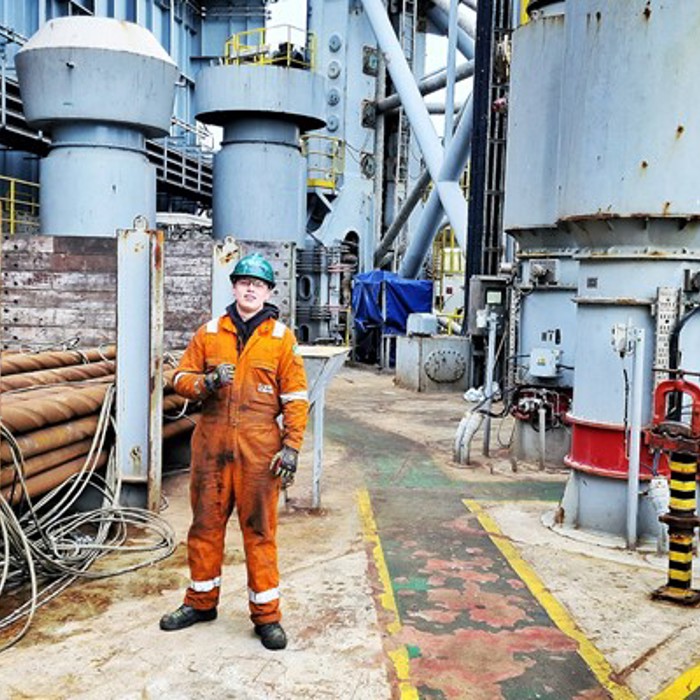 A worker in an orange jumpsuit stands on an industrial platform surrounded by large machinery and equipment.