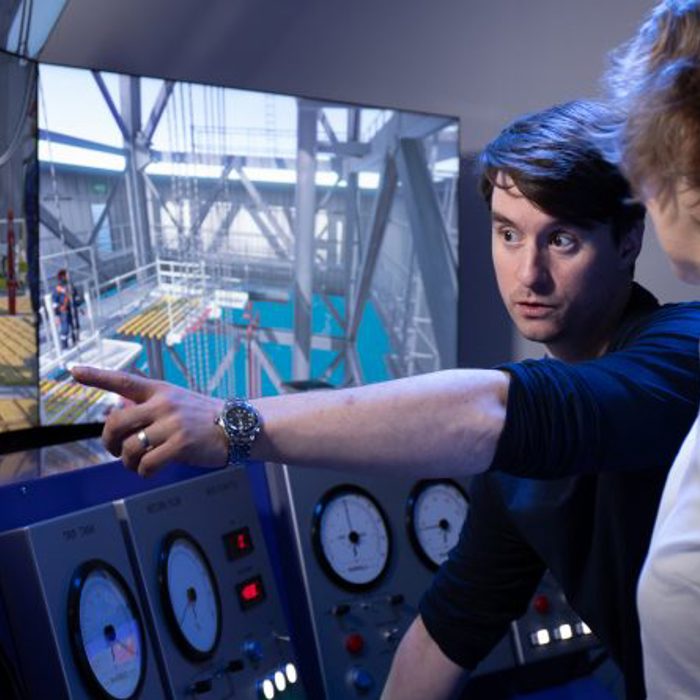 Person points at a screen displaying industrial controls and machinery simulations while explaining to another individual in front of control panels with analog gauges.