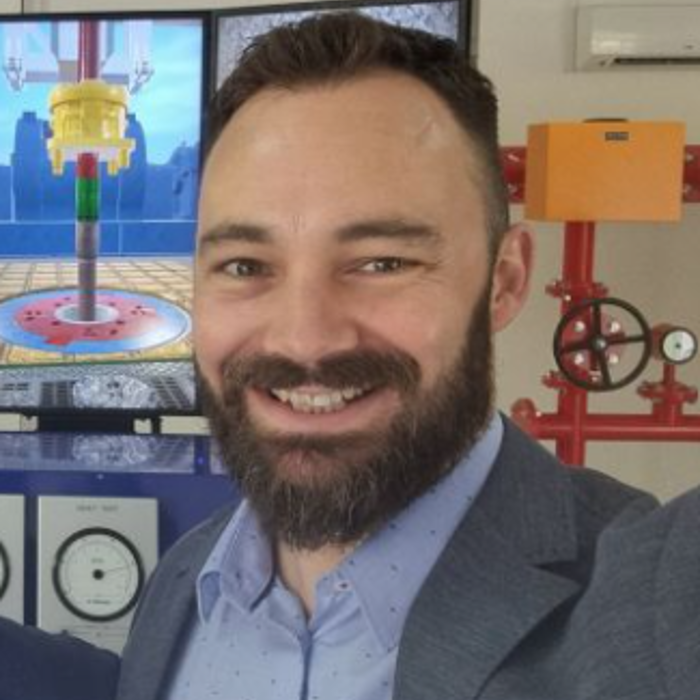 A man with a beard and short hair is smiling in front of a background with a television screen showing a brightly colored industrial scene and red industrial equipment.