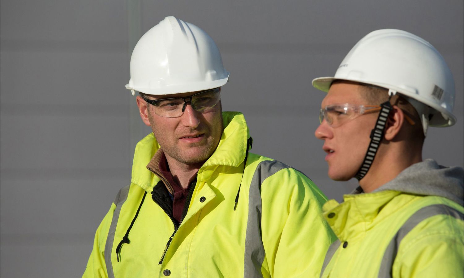 Two construction workers in high-visibility jackets and white helmets engage in conversation outside, discussing a recent training session.