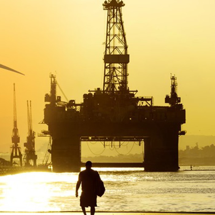 Silhouette of a person walking towards a large offshore oil rig at sunset, with wind turbines visible in the background.