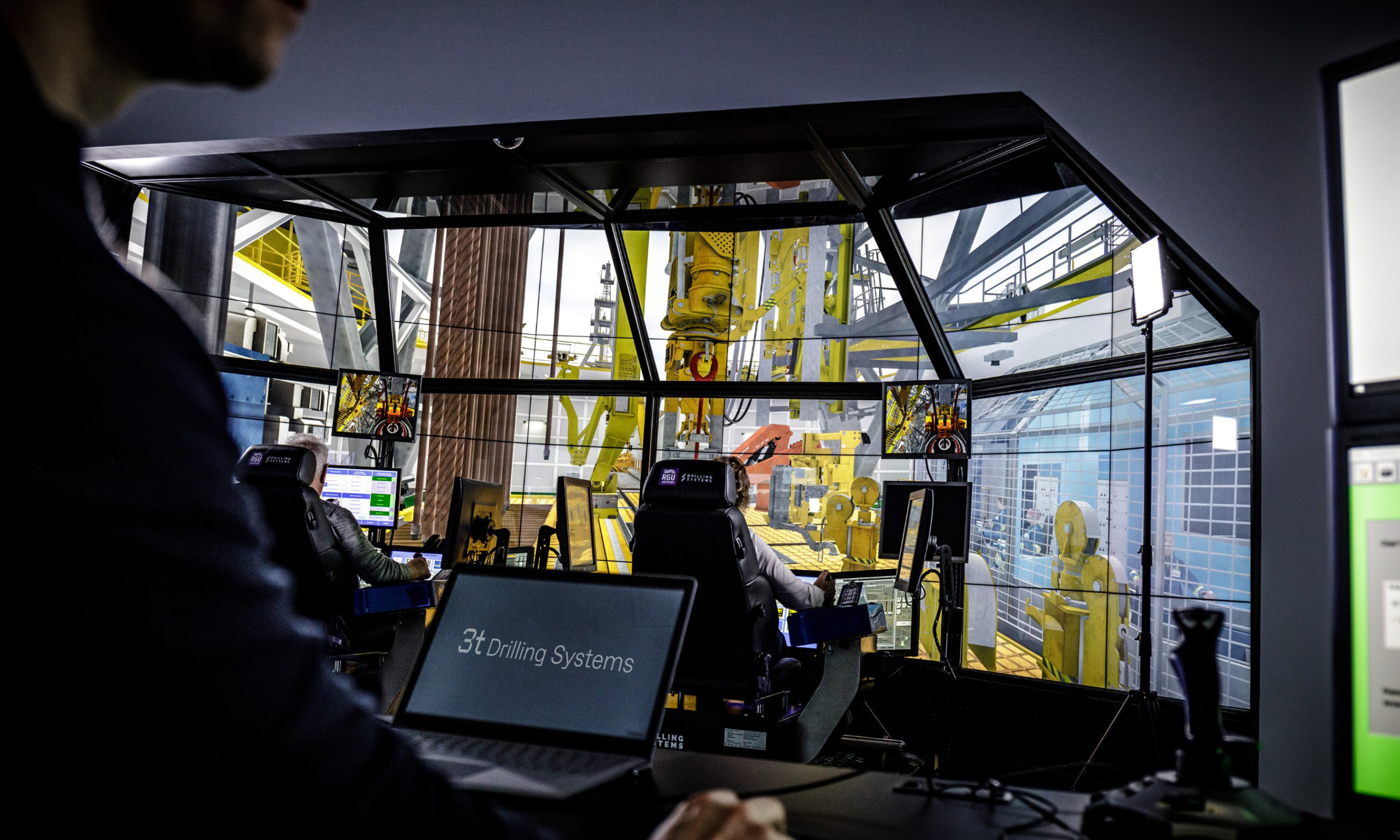 People operating a drilling simulator with multiple screens displaying various control panels and machinery. A person in the foreground is working on a laptop that says "3T Drilling Systems".
