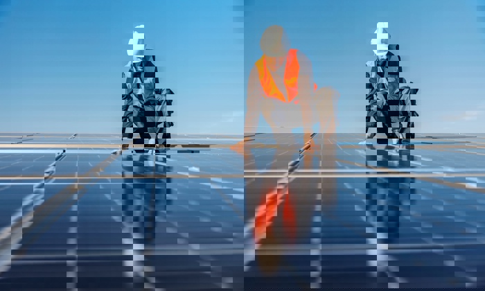 Worker in a safety vest and helmet installs solar panels on a sunny day.