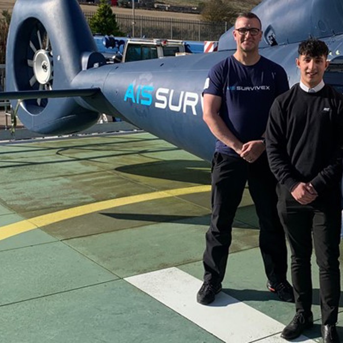 Three people standing on a helipad in front of a helicopter. The man on the left is wearing a navy blue shirt, the man in the middle is wearing a black outfit, and the woman on the right is in a navy blue jacket.
