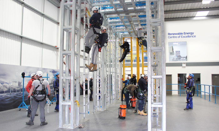 People wearing safety gear are practicing climbing and rescue techniques on metal structures inside an indoor training facility labeled "Renewable Energy Centre of Excellence.
