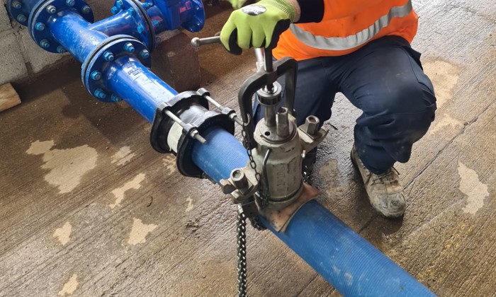 A worker in an orange safety jacket and gloves is using a wrench to repair a blue industrial pipe on a wet concrete floor, showcasing skills honed through extensive training.