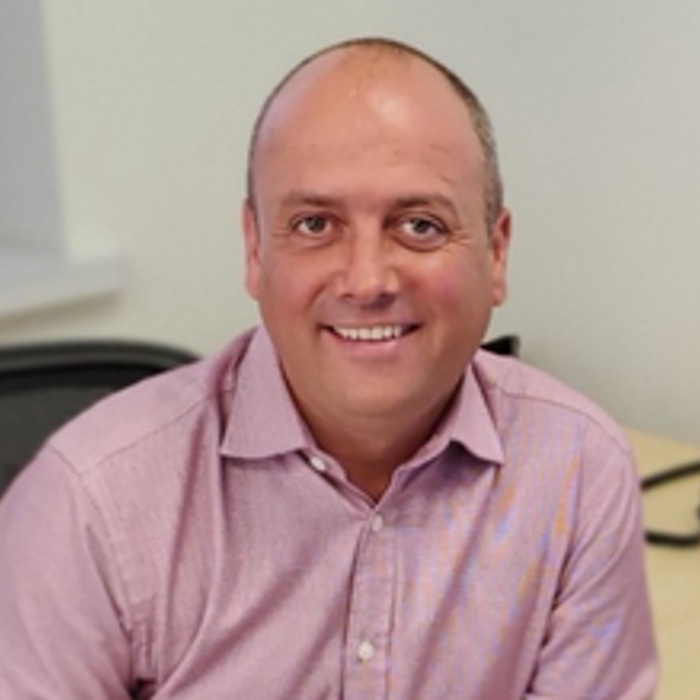 A man in a pink shirt sits at a desk with a phone and window in the background, smiling at the camera.