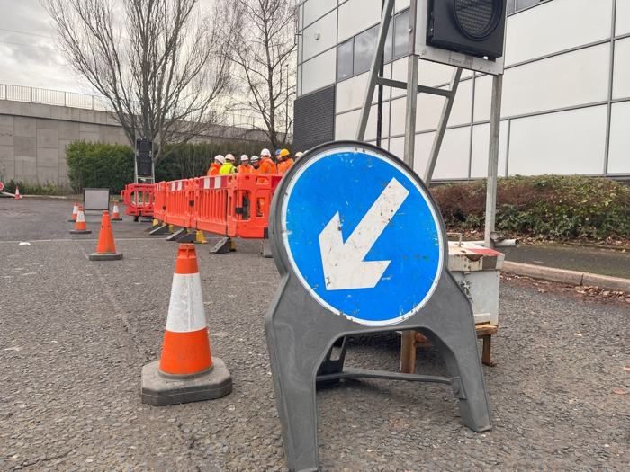 Traffic cones and a blue arrow sign direct to the right. Workers in orange vests, undergoing training, are near a row of red barriers next to a gray building.