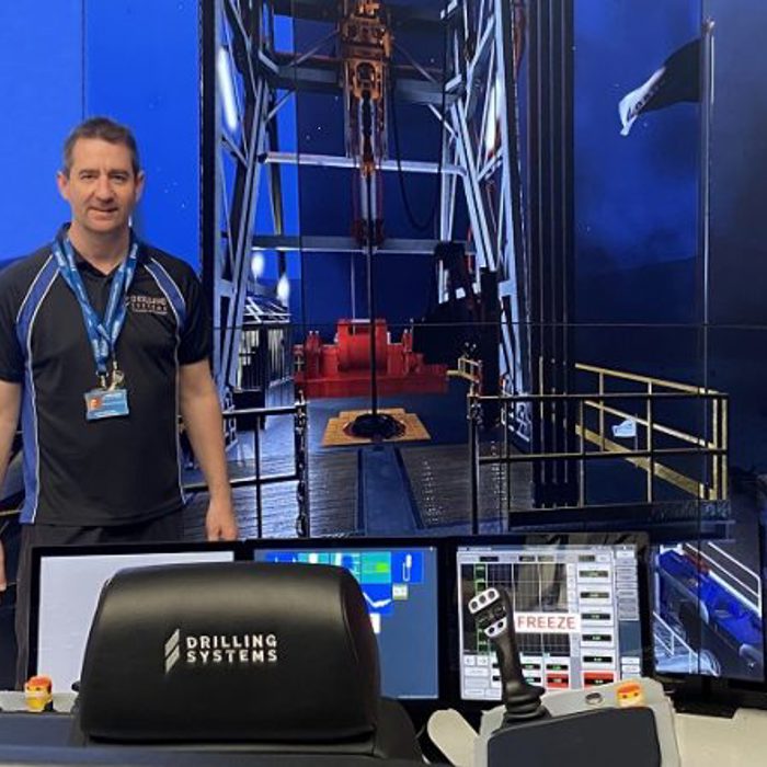 A man stands in front of a drilling simulation setup with multiple screens displaying drilling operations. He is wearing a blue shirt and a name badge. The area is well-lit with technical equipment visible.