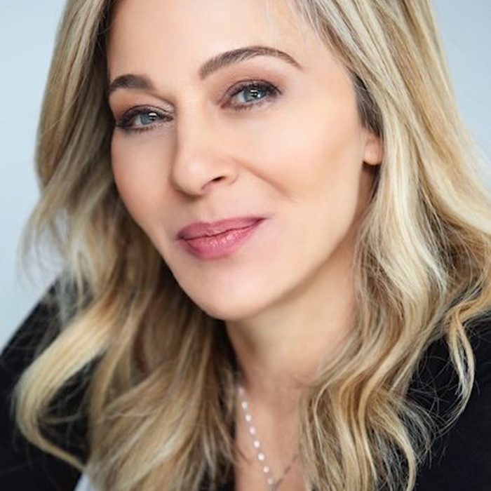 A woman with long, wavy blond hair and fair skin, wearing a black top, smiles softly.