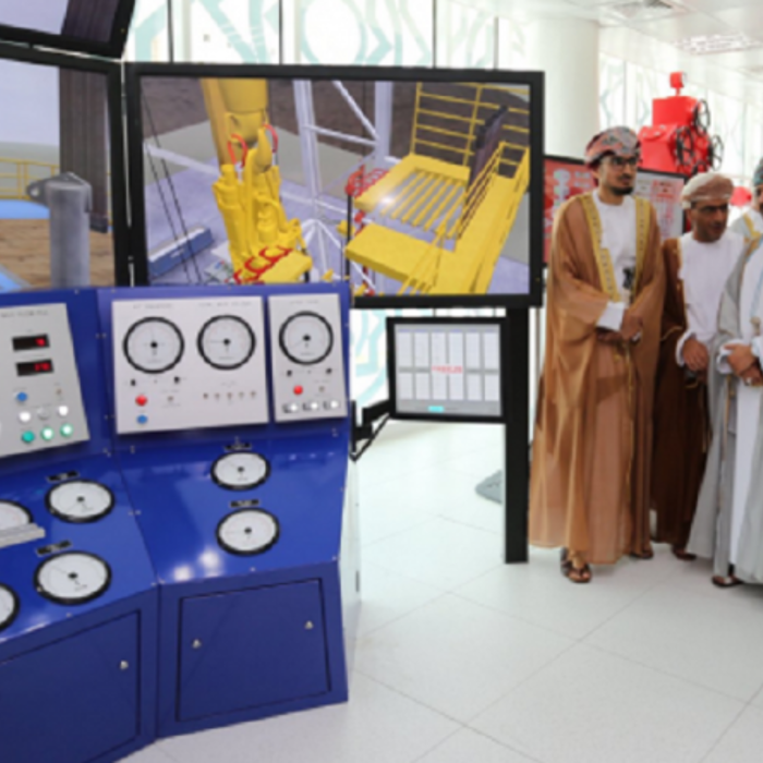 A group of men in traditional and Western attire gather around a control panel and display screens showing technical diagrams. One man in white points to the screens as he explains.