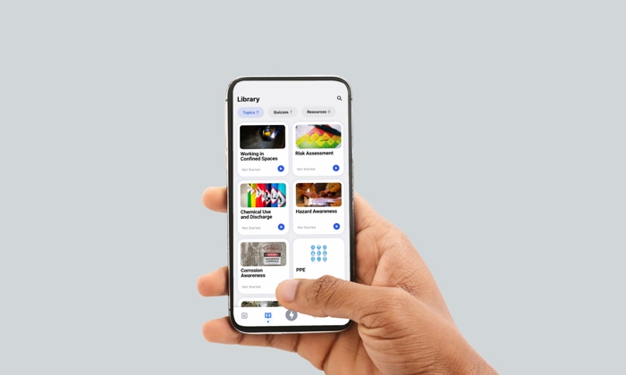 A hand holds a smartphone displaying a library of safety-related courses, including Working in Confined Spaces, Basic Assessment, and PPE, among others.