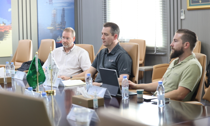 Four men seated at a conference table engage in a training meeting. Various items, including laptops and water bottles, are visible on the table. Flags are also present.