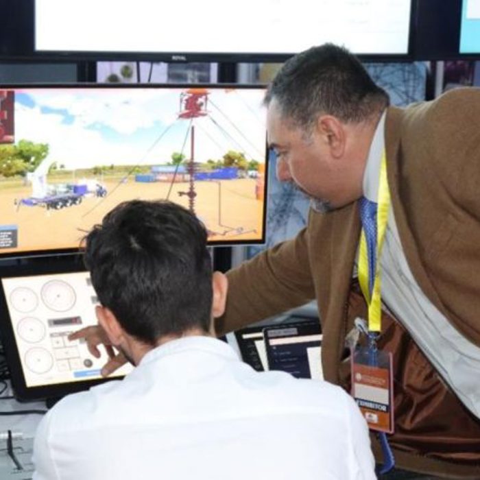 Two men interact with a control panel and monitor display in an industrial booth setting, demonstrating equipment operation.