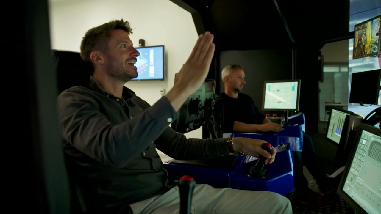Two men are operating a flight simulator in a control room, with one raising his hand while both focus on screens and controls in front of them.