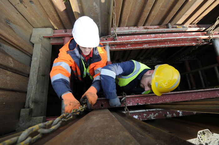 Two construction workers in safety gear are handling a metal chain inside a trench with surrounding wooden and metal supports, skillfully applying their training to ensure a safe and efficient process.