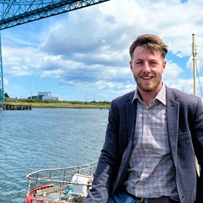 A man in a blazer stands on a boat with a river and a large blue industrial structure in the background under a partly cloudy sky.
