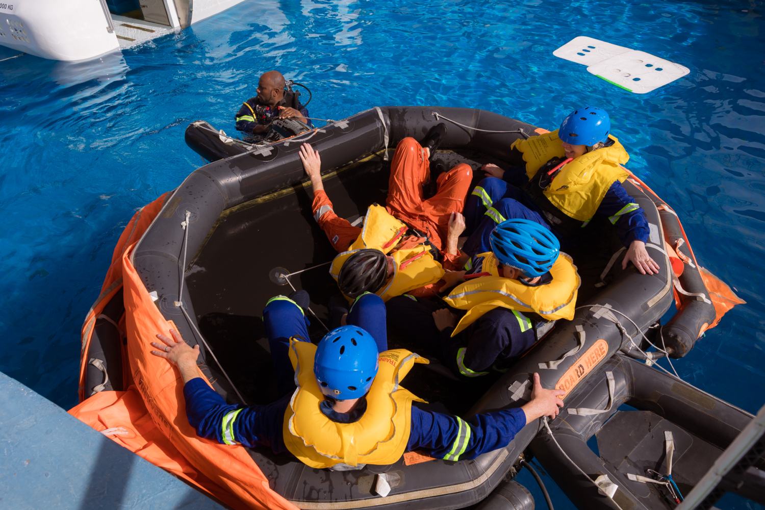 4 people sitting in black survival raft with safety diver in water