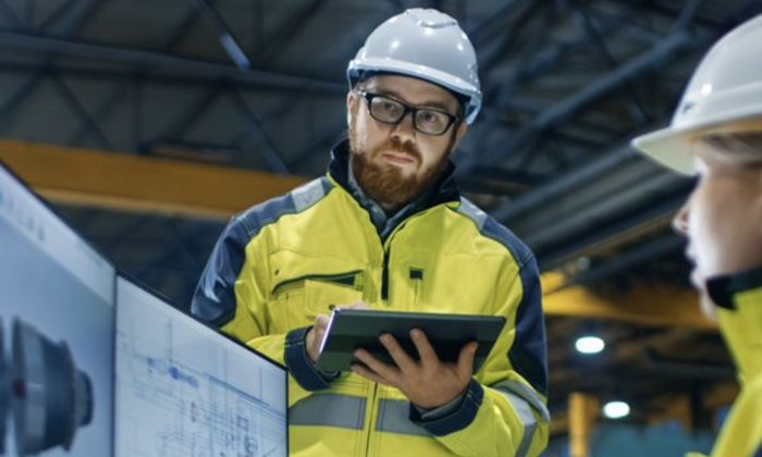 Two workers in high-visibility jackets and hard hats are reviewing information on a tablet and a large screen in an industrial setting.