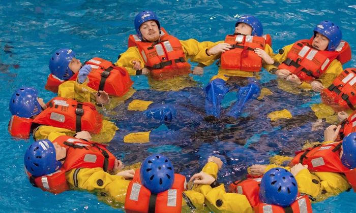 A group of people wearing orange life jackets and helmets form a circle while floating in a pool, participating in a training exercise.