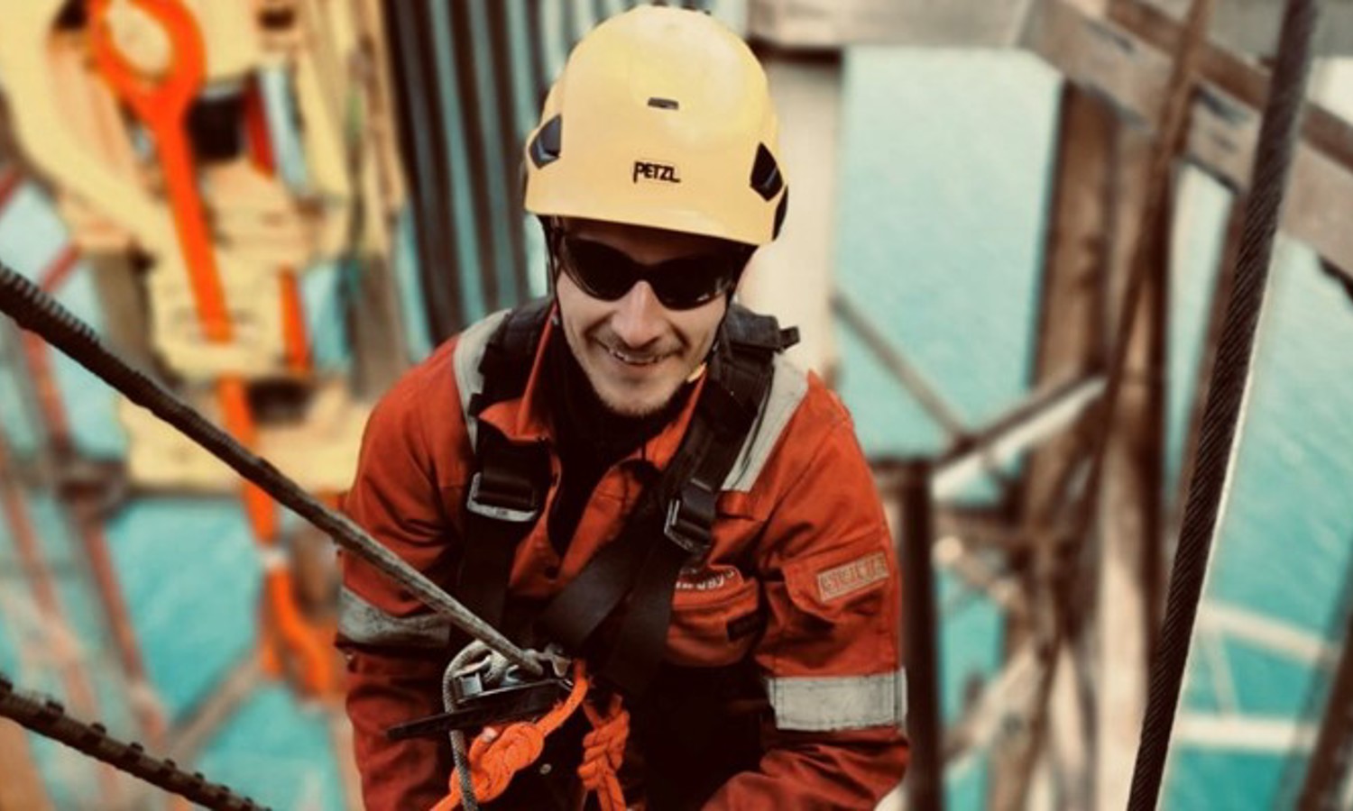 A worker wearing a yellow helmet and safety equipment is climbing a structure with ropes, with a large body of water visible in the background.