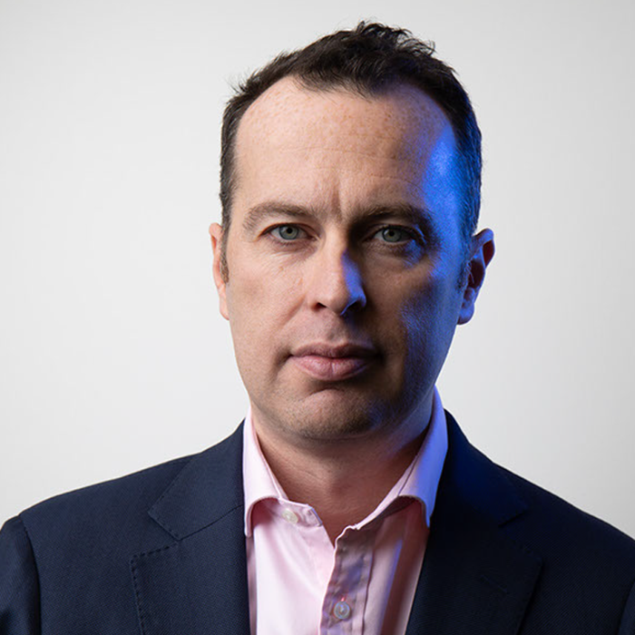A man with short dark hair, wearing a blue suit and a pink shirt, looks directly at the camera against a plain white background.